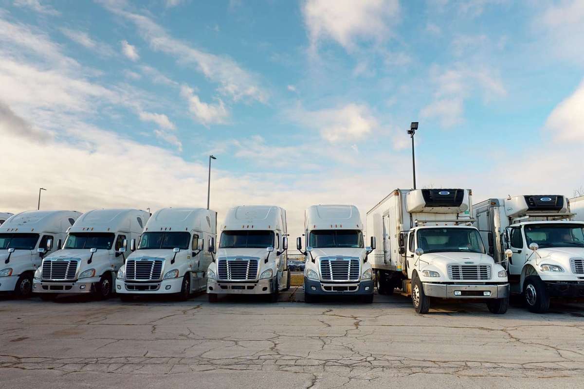 A line of white semi-trucks and box trucks parked in a lot against a blue cloudy sky.