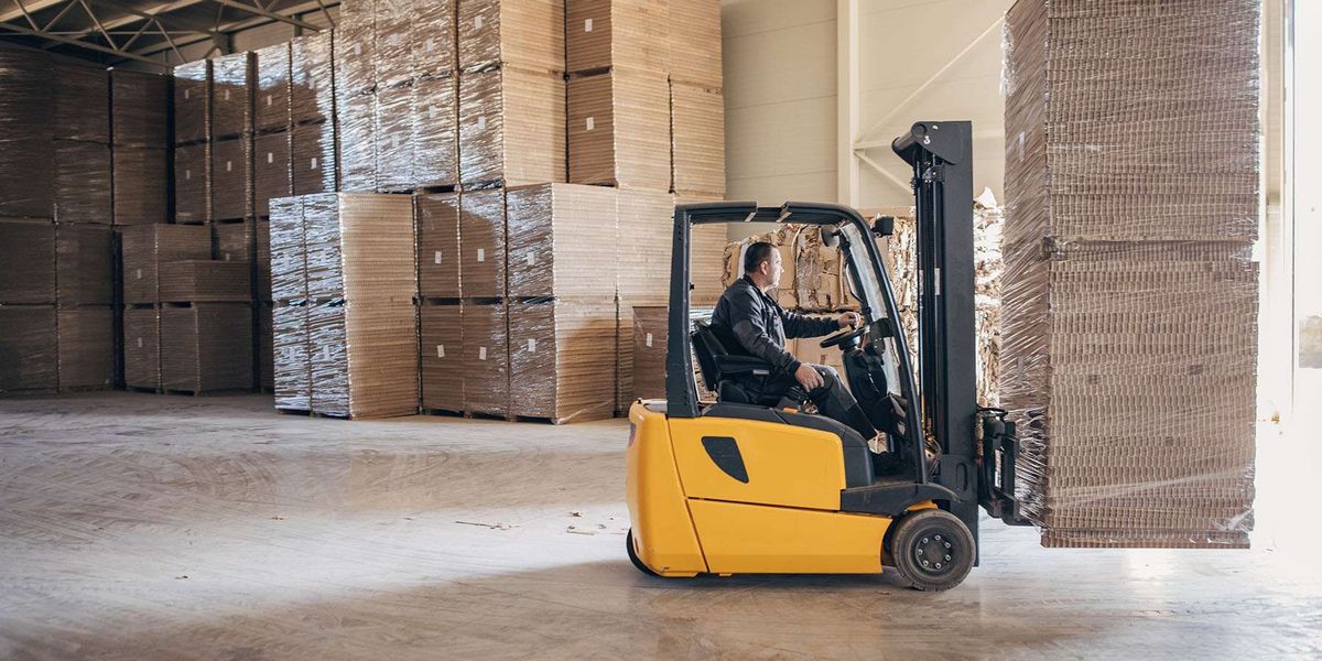 A man drives a yellow forklift carrying a stack of cardboard.