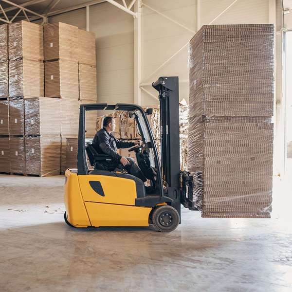 A man drives a yellow forklift carrying a stack of cardboard.
