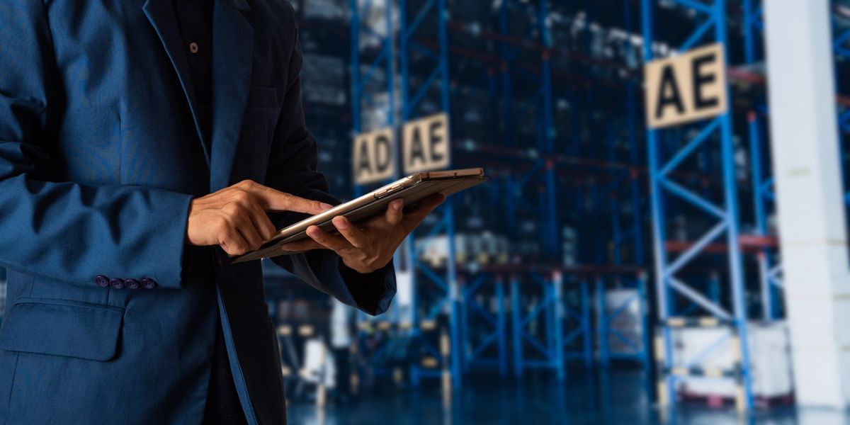 A man in a blue suit scrolls on a tablet while standing in a warehouse.