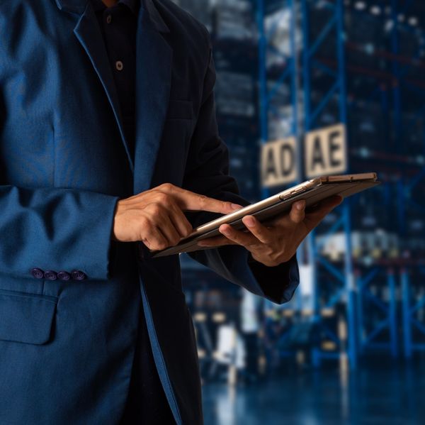 A man in a blue suit scrolls on a tablet while standing in a warehouse.