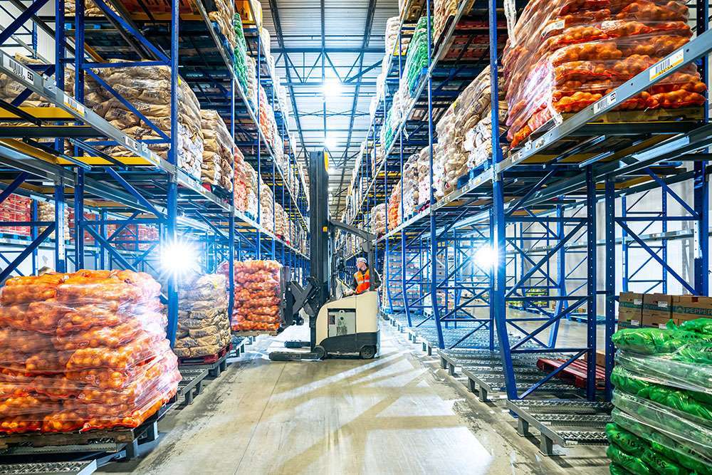A warehouse worker sits on a machine between two tall racks of food.