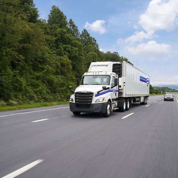A white Penske semi-truck drives up a highway next to green trees.