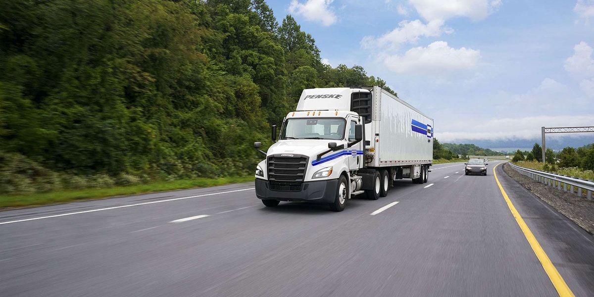 A white Penske semi-truck drives up a highway next to green trees.