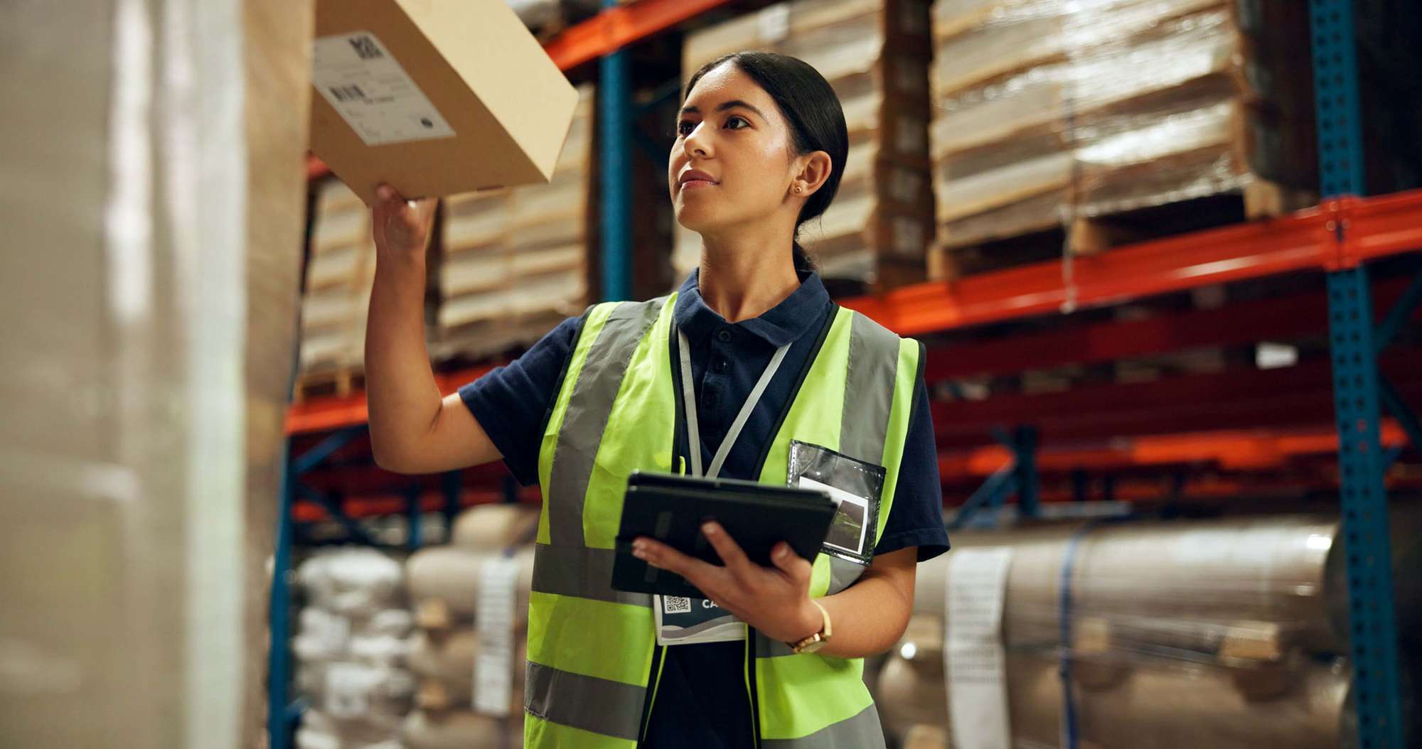 A woman in a neon vest holds an ipad while pulling a box from a warehouse shelf.