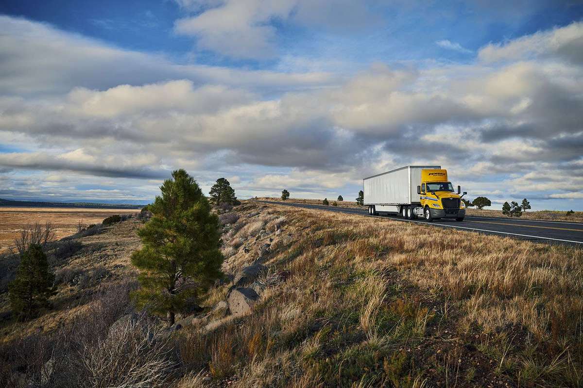 A yellow and white Penske semi-truck drives along a country road under a blue cloudy sky.