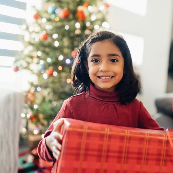 A young girl holds a gift wrapped in red wrapping paper with a lit Christmas tree behind her.