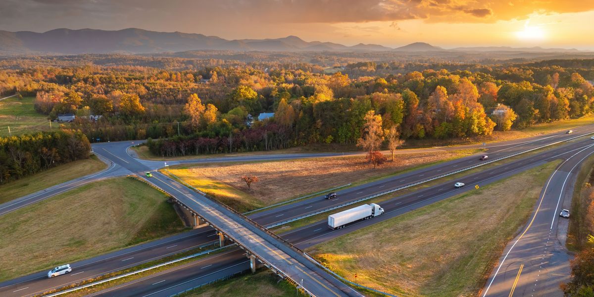 Aerial view of a white semi-truck and other cars drive up a freeway at sunset.
