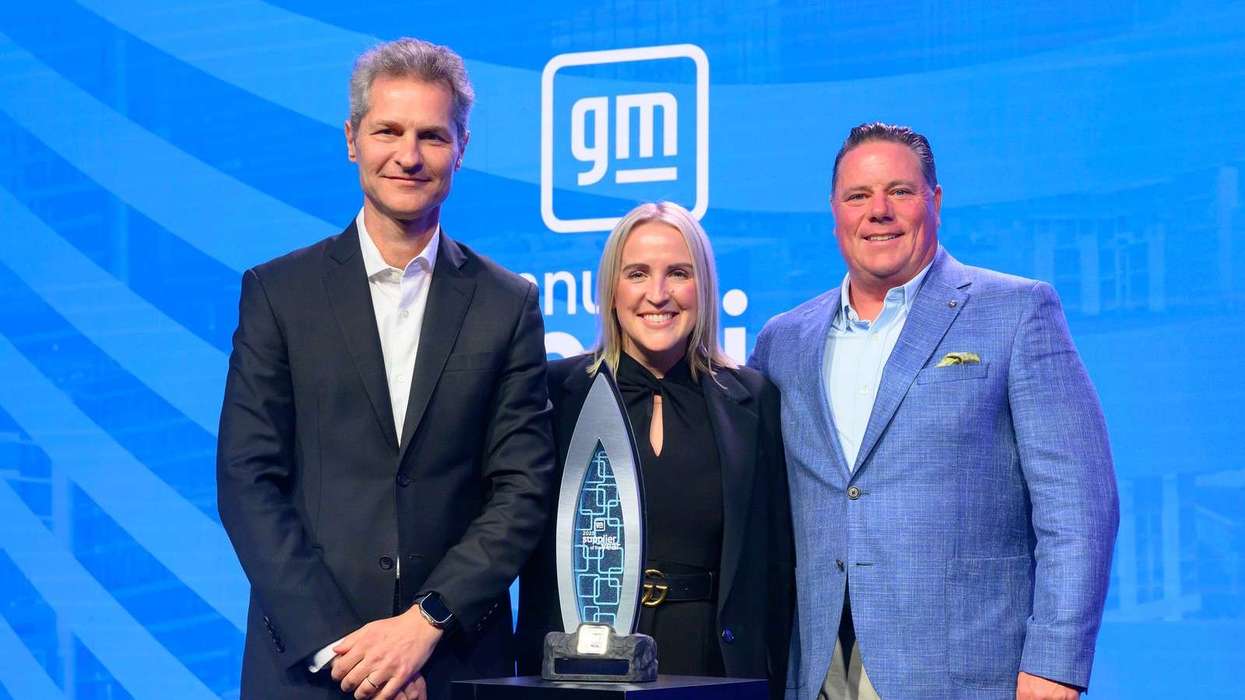 Three people stand amid a blue background around the GM Supplier of the Year Award Trophy