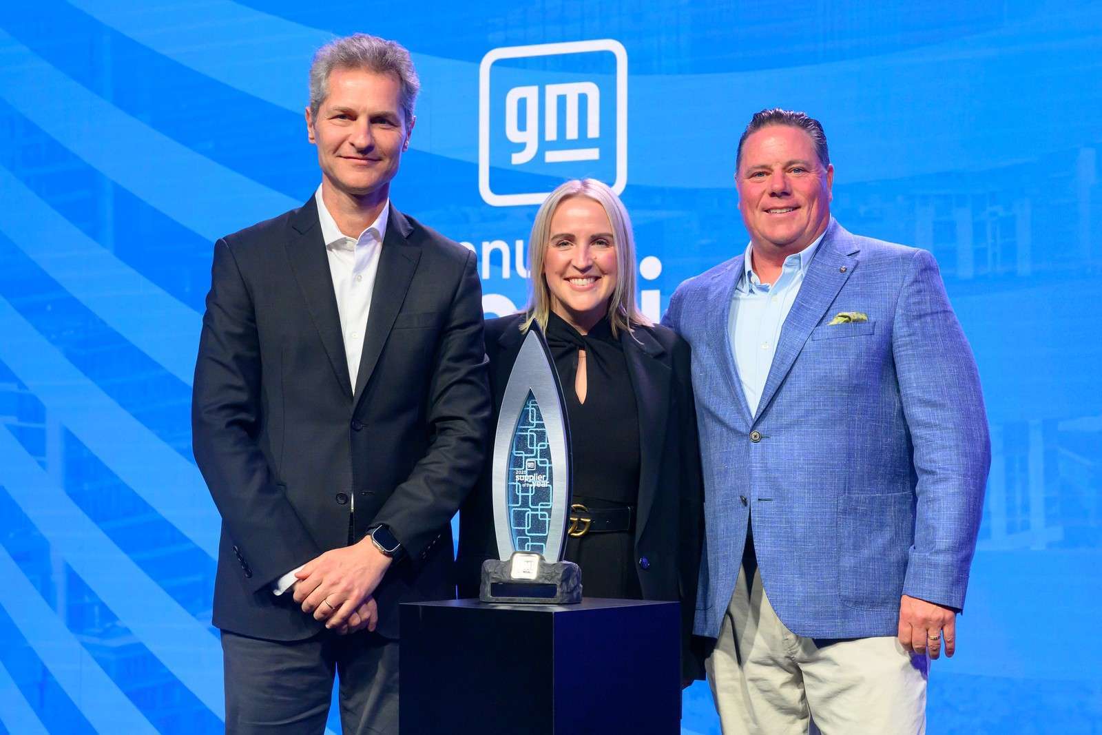 Three people stand amid a blue background around the GM Supplier of the Year Award Trophy