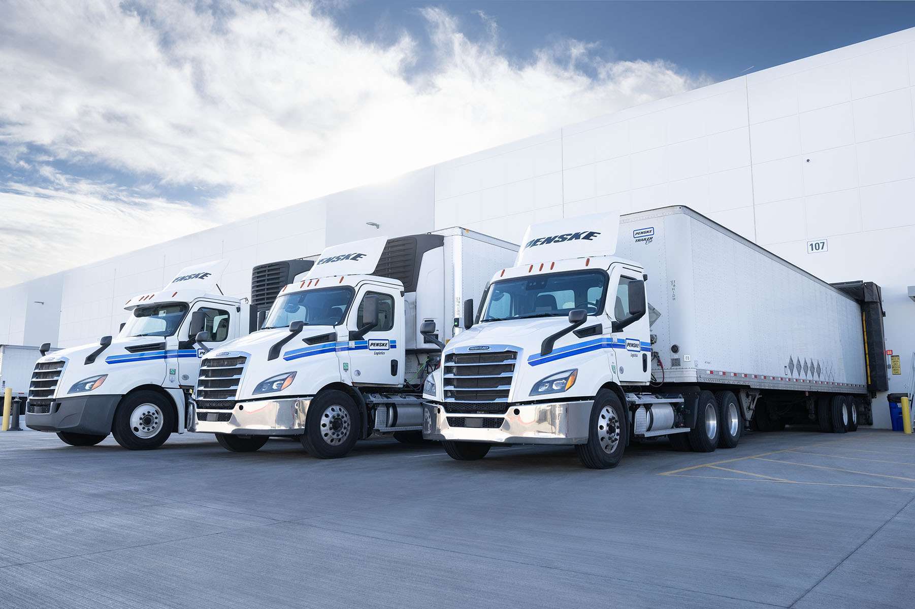 Three white Penske semi-trucks are backed into a warehouse loading dock.