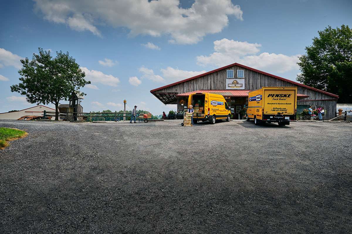 Two yellow Penske trucks unload boxes outside a brown barn farmer's market