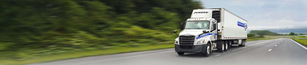 White freight truck driving on a highway, blurred green foliage in the background.